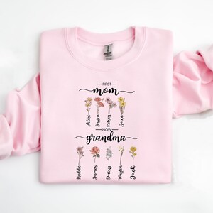 May include: Pink sweatshirt with a floral design and the text "First Mom - Now Grandma" with names listed below each flower.