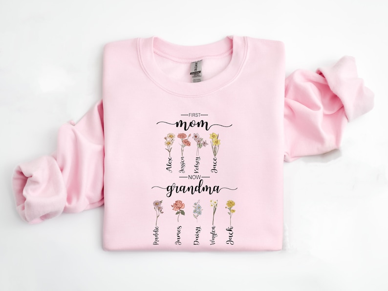 May include: Pink sweatshirt with a floral design and the text "First Mom - Now Grandma" with names listed below each flower.