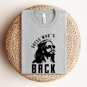 Puede incluir: Una camiseta gris con un gr&aacute;fico negro de Jes&uacute;s con gafas de sol y una corona de espinas. El texto "GUESS WHO'S BACK" est&aacute; impreso debajo del gr&aacute;fico.