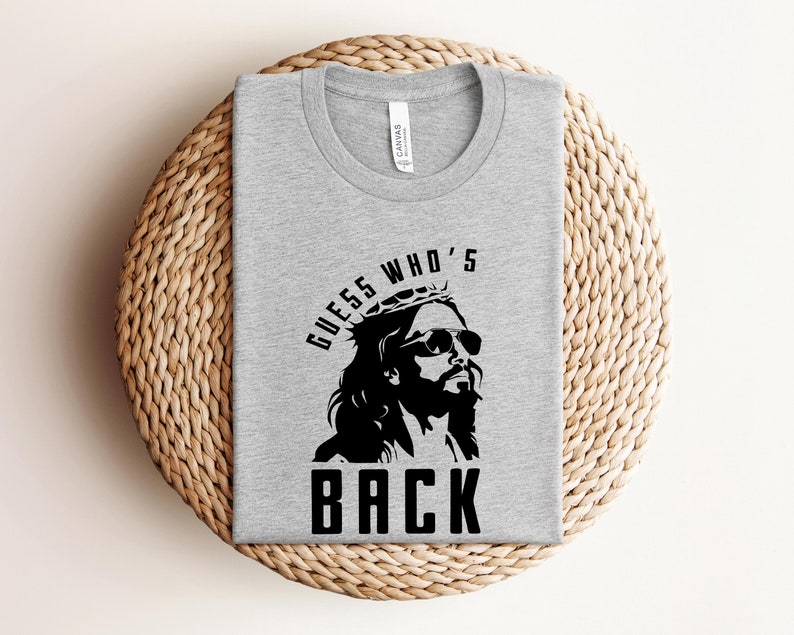 Puede incluir: Una camiseta gris con un gr&aacute;fico negro de Jes&uacute;s con gafas de sol y una corona de espinas. El texto "GUESS WHO'S BACK" est&aacute; impreso debajo del gr&aacute;fico.