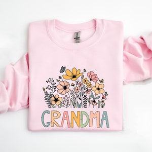 Puede incluir: Una sudadera rosa con un dise&ntilde;o floral y la palabra "GRANDMA" en letras de colores.