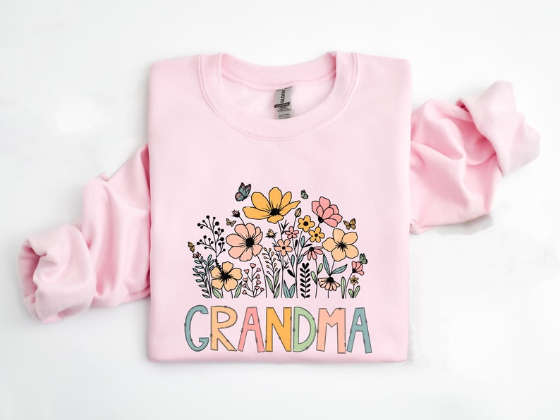 Puede incluir: Una sudadera rosa con un dise&ntilde;o floral y la palabra "GRANDMA" en letras de colores.