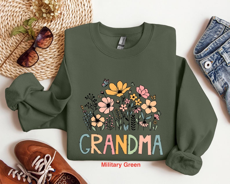 Puede incluir: Una sudadera verde con un dise&ntilde;o floral y la palabra "Grandma" en letras de colores. La sudadera est&aacute; sobre un fondo blanco.