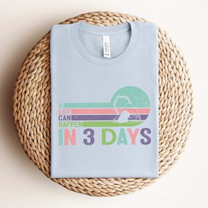 Puede incluir: Camiseta azul claro con un gr&aacute;fico de estilo retro que dice "A lot can happen in 3 days". El gr&aacute;fico presenta un dise&ntilde;o a rayas de colores con una ola y una palmera.