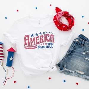 Puede incluir: Una camiseta blanca con un gr&aacute;fico rojo, blanco y azul que dice "America the Beautiful". La camiseta est&aacute; colocada sobre una superficie blanca con una bandana roja, pantalones cortos de mezclilla azul y confeti rojo y azul.