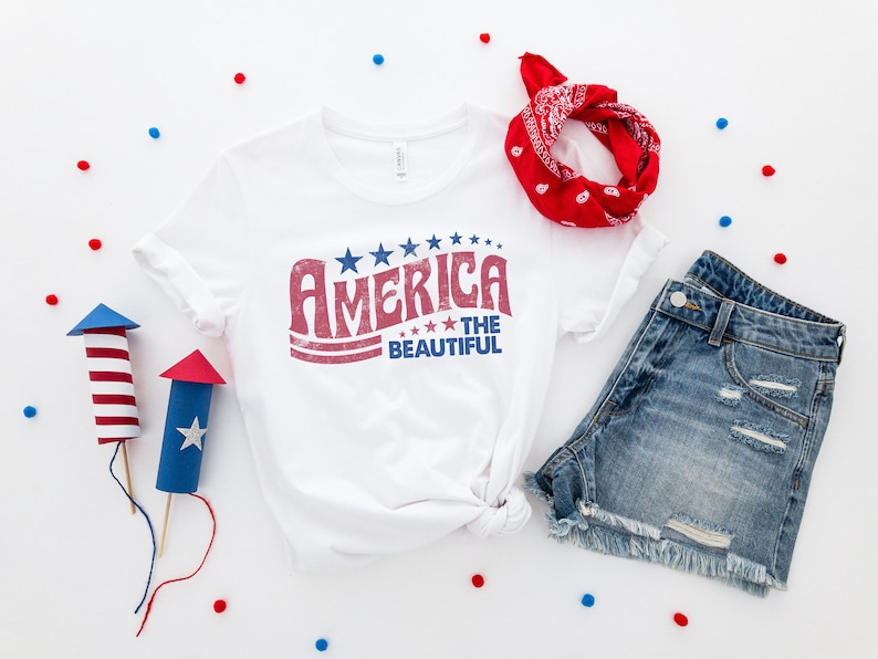 Puede incluir: Una camiseta blanca con un gr&aacute;fico rojo, blanco y azul que dice "America the Beautiful". La camiseta est&aacute; colocada sobre una superficie blanca con una bandana roja, pantalones cortos de mezclilla azul y confeti rojo y azul.