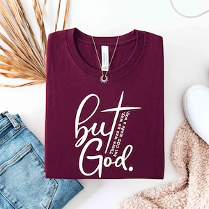 May include: A maroon t-shirt with the words "but God" in white script and a cross design. The text "There was no way, but GOD made a way" is also printed on the shirt. A silver necklace with a sunburst pendant is on the shirt.