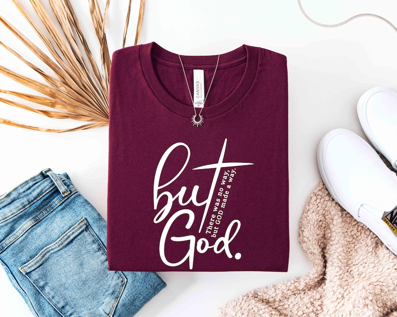 May include: A maroon t-shirt with the words "but God" in white script and a cross design. The text "There was no way, but GOD made a way" is also printed on the shirt. A silver necklace with a sunburst pendant is on the shirt.