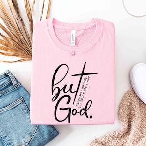 May include: A light pink t-shirt with the words "but God" in a black script font, with a cross graphic. The text "There was no way, but GOD made a way" is also printed on the shirt. The shirt is folded and placed next to blue jeans and white shoes.