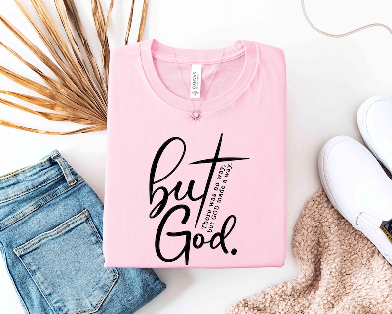 May include: A light pink t-shirt with the words "but God" in a black script font, with a cross graphic. The text "There was no way, but GOD made a way" is also printed on the shirt. The shirt is folded and placed next to blue jeans and white shoes.