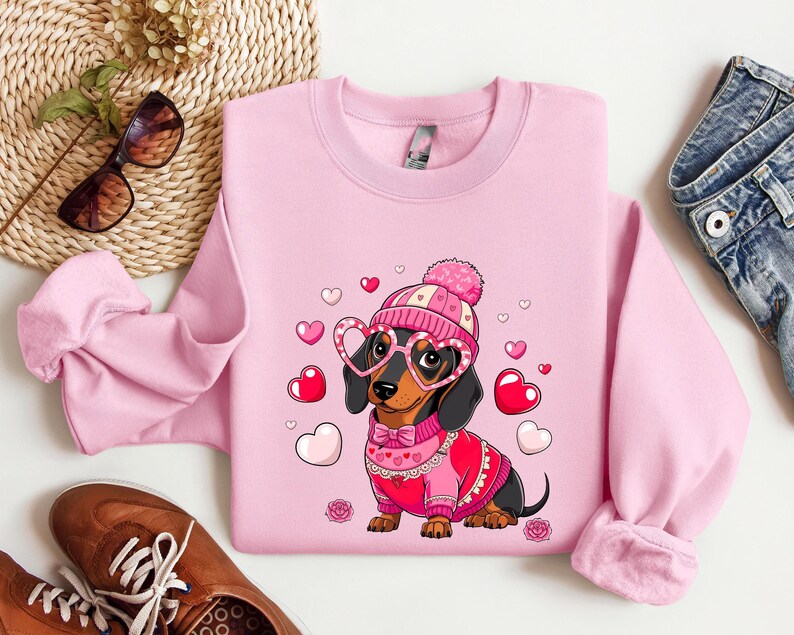 May include: Pink sweatshirt with a cartoon dachshund wearing a pink and white hat and heart-shaped glasses. The dachshund is wearing a red sweater with a white collar and a bow tie. The sweatshirt has a Valentine's Day theme with hearts scattered around the dachshund.