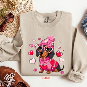 May include: A beige sweatshirt with a graphic of a dachshund wearing a pink hat, pink heart-shaped glasses, and a pink sweater with a heart pattern. The dachshund is surrounded by pink hearts. The text "SAND" is printed on the bottom of the sweatshirt.