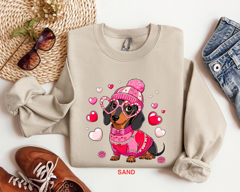 May include: A beige sweatshirt with a graphic of a dachshund wearing a pink hat, pink heart-shaped glasses, and a pink sweater with a heart pattern. The dachshund is surrounded by pink hearts. The text "SAND" is printed on the bottom of the sweatshirt.