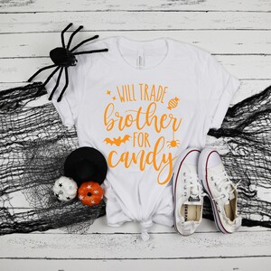 Puede incluir: Camiseta blanca con texto naranja "Will Trade Brother For Candy" y gr&aacute;ficos de Halloween. La camiseta se combina con zapatillas blancas, una ara&ntilde;a negra y calabazas decorativas.