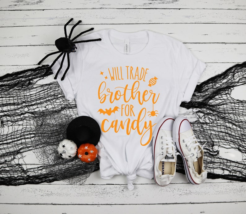 Puede incluir: Camiseta blanca con texto naranja "Will Trade Brother For Candy" y gr&aacute;ficos de Halloween. La camiseta se combina con zapatillas blancas, una ara&ntilde;a negra y calabazas decorativas.