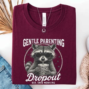 May include: Burgundy t-shirt featuring a raccoon wearing sunglasses and drinking from a leopard-print cup. The shirt has the text "GENTLE PARENTING Dropout EST. THIS MORNING" in white. A silver necklace with a sun pendant is also visible.