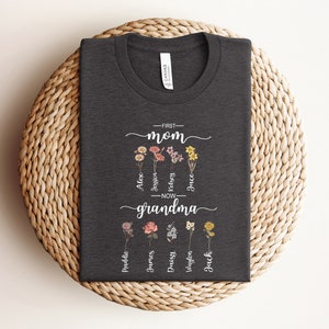 May include: A dark gray t-shirt with the text "First Mom - Now Grandma" and a floral design. The names Alex, Jessin, Kelsey, Jace,  Madeline, James, Daisy,  Waylon, and Jack are written around the flowers.