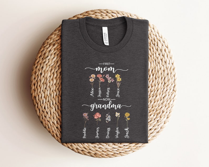 May include: A dark gray t-shirt with the text "First Mom - Now Grandma" and a floral design. The names Alex, Jessin, Kelsey, Jace,  Madeline, James, Daisy,  Waylon, and Jack are written around the flowers.