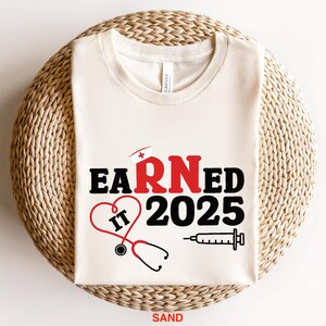May include: A white t-shirt with the text "EARNED IT 2025" in black and red. The "RN" is in red and has a small red cross on top. A red heart with the text "IT" inside is next to the text. A stethoscope and a syringe are below the text.
