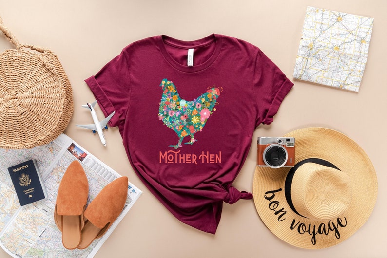 Puede incluir: Camiseta burdeos con un dise&ntilde;o floral de gallina y el texto "MOTHER HEN". La imagen incluye accesorios de viaje como un bolso de paja, pasaporte, mapa, avi&oacute;n, c&aacute;mara y un sombrero de sol con el texto "bon voyage".