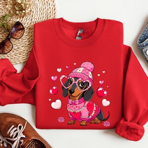 May include: Red sweatshirt with a cartoon dachshund wearing a pink hat and heart-shaped glasses. The dachshund is wearing a pink sweater with a white collar and a bow tie. The sweatshirt has a Valentine's Day theme with pink hearts scattered around the dachshund.
