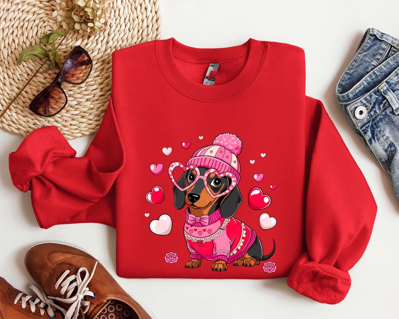 May include: Red sweatshirt with a cartoon dachshund wearing a pink hat and heart-shaped glasses. The dachshund is wearing a pink sweater with a white collar and a bow tie. The sweatshirt has a Valentine's Day theme with pink hearts scattered around the dachshund.