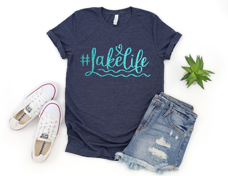 May include: A navy blue t-shirt with a turquoise glitter design that says "#lakelife" with a small heart above the word "life". The shirt is paired with distressed denim shorts and white sneakers.