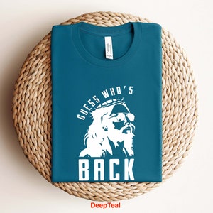 Puede incluir: Una camiseta azul turquesa con un gr&aacute;fico blanco de una persona con una corona y gafas de sol. El texto "Guess Who's Back" est&aacute; impreso en la camiseta.