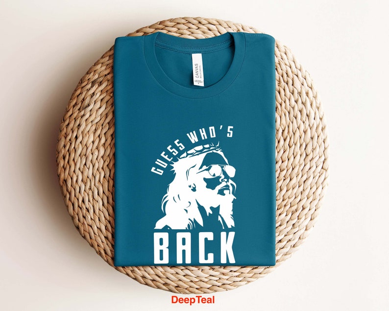 Puede incluir: Una camiseta azul turquesa con un gr&aacute;fico blanco de una persona con una corona y gafas de sol. El texto "Guess Who's Back" est&aacute; impreso en la camiseta.