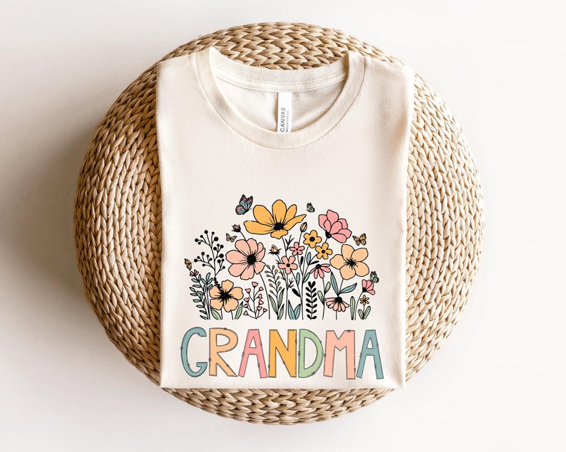 Puede incluir: Una camiseta blanca con un dise&ntilde;o floral y la palabra "GRANDMA" en letras de colores.