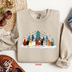 May include: A beige sweatshirt with a Christmas-themed design featuring a group of chickens wearing Santa hats and standing in front of a decorated Christmas tree. The text "SAND" is printed on the bottom of the sweatshirt.