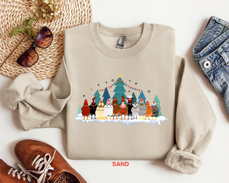 May include: A beige sweatshirt with a Christmas-themed design featuring a group of chickens wearing Santa hats and standing in front of a decorated Christmas tree. The text "SAND" is printed on the bottom of the sweatshirt.