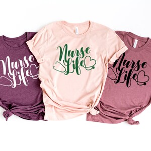 K&ouml;nnte beinhalten: Drei T-Shirts mit dem Text "Nurse Life" in einer kursiven Schrift. Die Shirts sind in verschiedenen Farben: weinrot, rosa und hellrosa. Jedes Shirt hat ein Stethoskop und ein Herz-Motiv.