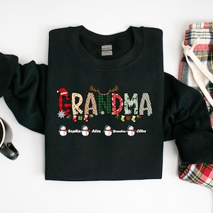 May include: Black sweatshirt with the word "GRANDMA" spelled out in colorful patterned letters. The letters are decorated with Christmas-themed designs, including reindeer, snowflakes, and stockings. Below the word "GRANDMA" are four snowmen with the names "Sophia", "Alex", "Brandon", and "Olive" written underneath.