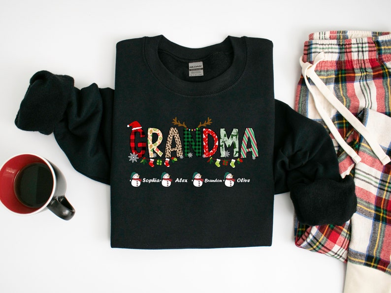 May include: Black sweatshirt with the word "GRANDMA" spelled out in colorful patterned letters. The letters are decorated with Christmas-themed designs, including reindeer, snowflakes, and stockings. Below the word "GRANDMA" are four snowmen with the names "Sophia", "Alex", "Brandon", and "Olive" written underneath.
