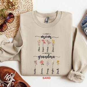 May include: A beige sweatshirt with the text "First Mom Now Grandma" and a floral design. The text is in black and the flowers are in various colors. The sweatshirt has a crew neck and long sleeves.
