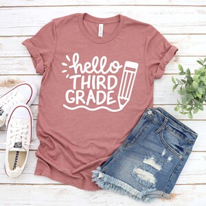 Op de afbeelding: Een stoffig roze T-shirt met de tekst "hello third grade" en een potloodgrafiek in wit.