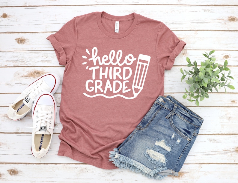 Op de afbeelding: Een stoffig roze T-shirt met de tekst "hello third grade" en een potloodgrafiek in wit.