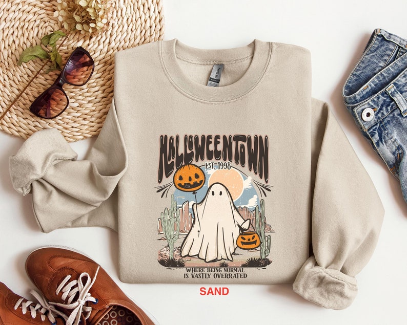 May include: A beige sweatshirt with a graphic print of a ghost holding a jack-o-lantern. The graphic says "Halloweentown est. 1998" and "Where being normal is vastly overrated SAND".