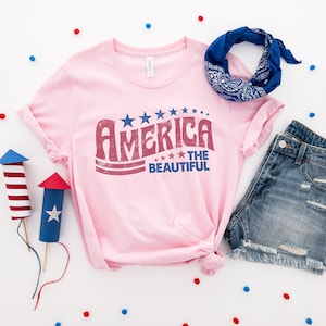 Puede incluir: Una camiseta rosa con un gr&aacute;fico de estilo vintage que dice "America the Beautiful" en letras rojas y azules con estrellas. La camiseta est&aacute; colocada sobre una superficie blanca con confeti azul y rojo, un pa&ntilde;uelo azul y unos pantalones cortos de mezclilla.