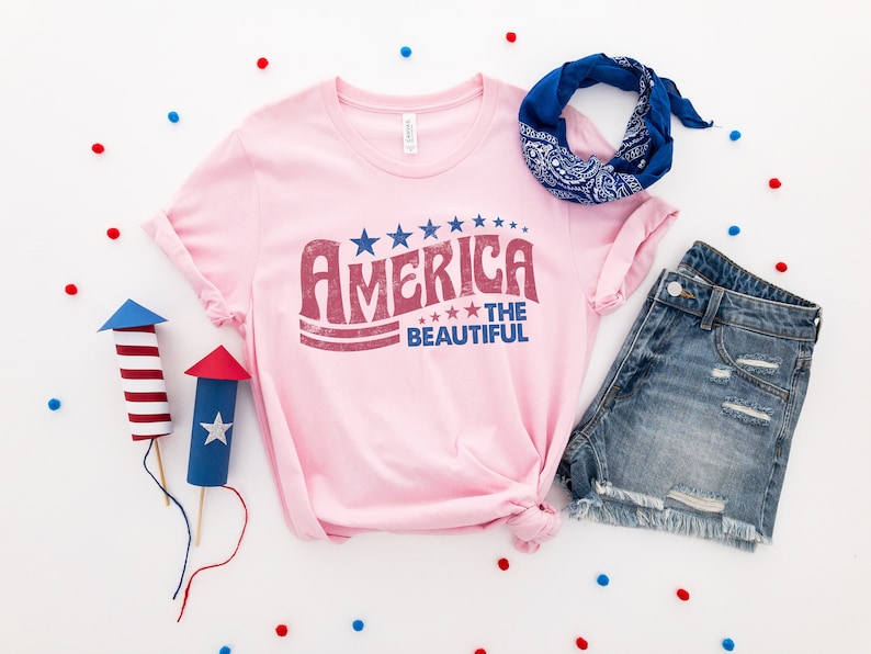 Puede incluir: Una camiseta rosa con un gr&aacute;fico de estilo vintage que dice "America the Beautiful" en letras rojas y azules con estrellas. La camiseta est&aacute; colocada sobre una superficie blanca con confeti azul y rojo, un pa&ntilde;uelo azul y unos pantalones cortos de mezclilla.