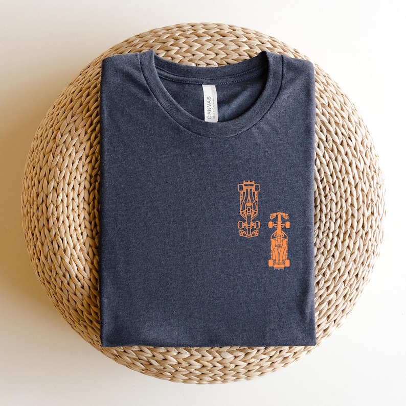 Peut inclure: Un t-shirt gris fonc&eacute; chin&eacute; avec un contour orange de deux voitures de course. Le t-shirt est sur un tapis de paille tiss&eacute;.