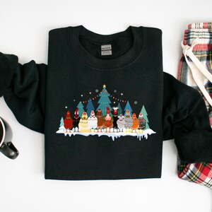 May include: Black sweatshirt with a Christmas-themed design featuring a group of chickens standing in front of a decorated Christmas tree. The chickens are wearing festive hats and scarves.