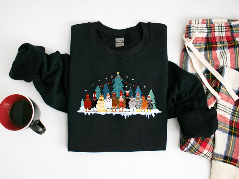 May include: Black sweatshirt with a Christmas-themed design featuring a group of chickens standing in front of a decorated Christmas tree. The chickens are wearing festive hats and scarves.