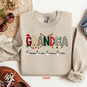 May include: A beige sweatshirt with a Christmas-themed "Grandma" graphic featuring a red hat, plaid, leopard print, and green and red stripes. The graphic is surrounded by a garland of holly, ornaments, and stockings. The names "Sophia", "Alex", "Brandon", and "Olive" are printed below the graphic. The sweatshirt is on a white background.