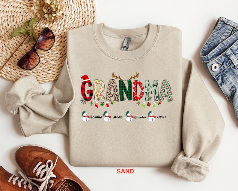 May include: A beige sweatshirt with a Christmas-themed "Grandma" graphic featuring a red hat, plaid, leopard print, and green and red stripes. The graphic is surrounded by a garland of holly, ornaments, and stockings. The names "Sophia", "Alex", "Brandon", and "Olive" are printed below the graphic. The sweatshirt is on a white background.