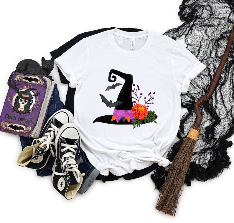 May include: White t-shirt with a black witch hat with purple and pink accents and floral details. The hat is surrounded by bats and has the text "Dark magic" on the bottom.