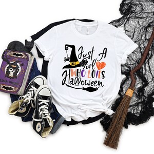 May include: White t-shirt with a black witch hat, spider, and the text "Just a girl who loves Halloween".