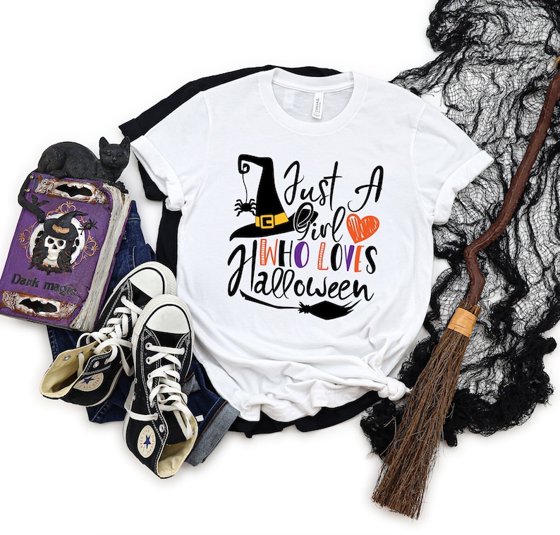 May include: White t-shirt with a black witch hat, spider, and the text "Just a girl who loves Halloween".