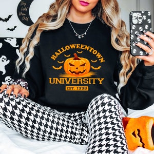 May include: Black sweatshirt with the text "Halloweentown University Est. 1998" and a cartoon orange pumpkin with a scary face. The sweatshirt is worn by a person with long blonde hair and is sitting on a white surface.